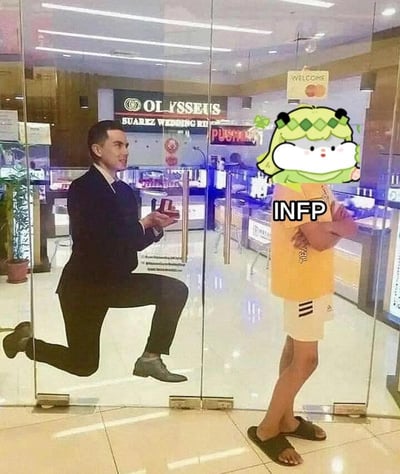 这是一张利用视觉错位制作的MBTI搞笑梗图，珠宝店的玻璃门内侧贴着男士单膝跪地举戒指求婚的广告海报，玻璃门外站着一位穿黄色T恤、白短裤、黑拖鞋的路人，路人头部被替换为绿色呆萌卡通表情包，胸前标注了MBTI人格类型“INFP”的字样，从画面视角看恰好形成了海报里的男士隔着玻璃向INFP路人求婚的错觉，错位效果充满笑点。