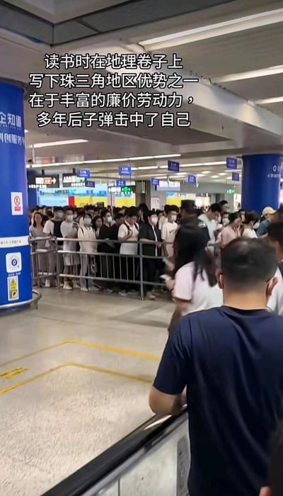 这是一张竖屏格式的原始实拍图，画面呈现的是珠三角某地铁站的通勤场景，自动扶梯出口处，大量戴着口罩的通勤人群被金属护栏引导着排队，人群密集，透着通勤时段的拥挤感。画面上方配有自嘲的文字：“读书时在地理卷子上写下珠三角地区优势之一在于丰富的廉价劳动力，多年后子弹击中了自己”，用一种戏谑的方式，表达了如今自己成为了当年地理题里描述的“廉价劳动力”的无奈与自嘲，精准戳中了在珠三角打拼的打工人的职场共鸣。