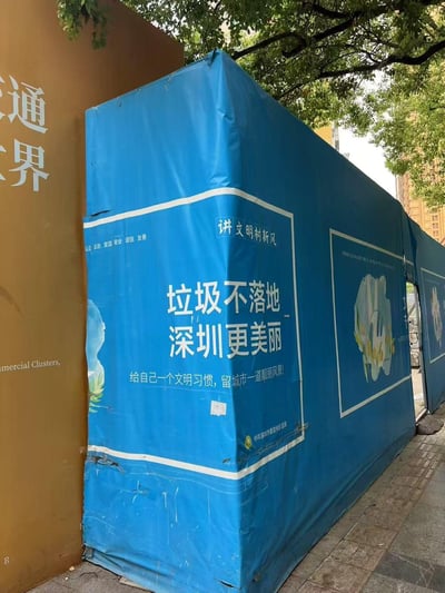 这是一张城市户外实拍照片，展示了蓝色的街道隔离围挡，上面印有深圳文明城市创建的宣传内容：
- 顶部标注「讲文明树新风」，旁侧印有社会主义核心价值观「公正 法治 爱国 敬业 诚信 友善」
- 核心标语为「垃圾不落地 深圳更美丽」
- 下方配有引导语「给自己一个文明习惯，留城市一道靓丽风景!」
- 底部标注内容由中共深圳市委宣传部监制
围挡旁侧是带有「通世界」字样的棕色墙面，上方有行道树，背景可见居民楼，呈现出日常的城市街道场景。