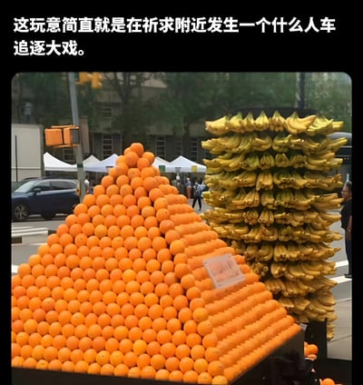 图片展示了一个街头水果摊，左侧是一座由大量橙子堆叠成的金字塔形果堆，右侧则是高耸的香蕉堆，水果摆放得极为密集且高耸。背景中可见城市街道、交通信号灯、行人和停放的车辆，呈现出典型的城市市集场景。图片上方配有文字“这玩意简直就是在祈求附近发生一个什么人车追逐大戏”，以幽默调侃的语气暗示水果堆叠得过于不稳定，仿佛随时会因意外（如人车追逐）而倒塌。