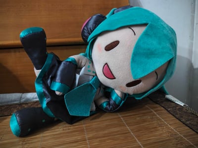 这张图展示了一个还原虚拟歌手初音未来形象的毛绒玩偶，它侧躺在竹制平面上，双眼弯成月牙状，嘴巴大张露出开心的笑容，姿态放松慵懒。玩偶完美复刻了初音未来标志性的青绿色发型、领带，搭配黑绿配色的服饰细节，整体造型可爱治愈，传递出满满的愉悦感。