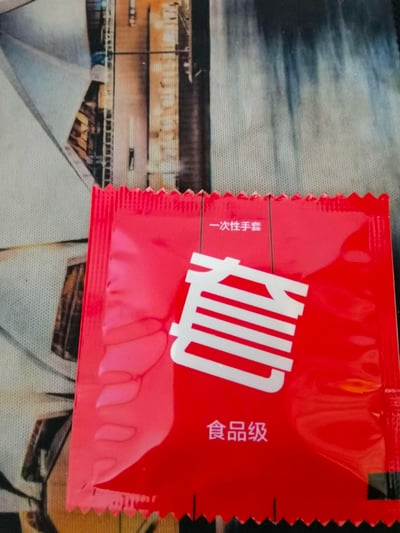 这是一张原始拍摄的图片，主体是一个红色独立包装的食品级一次性手套。包装设计风格类似安全套的外包装，中间印有超大号的白色“套”字，上方标注“一次性手套”，下方标注“食品级”。

这个包装利用了视觉联想的梗，通过类似安全套的包装样式和醒目的“套”字，制造出视觉反差感，容易让人第一眼误解为安全套，仔细查看文字才发现是一次性手套，充满趣味性。