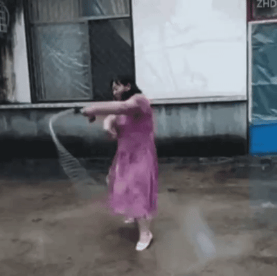 一位身穿粉色连衣裙的女子在户外场地表演甩鞭。她站在一栋老旧建筑前的泥土地面上，双手持鞭做出一系列流畅动作，包括双臂伸展、挥鞭转圈等。整个表演展现出她对鞭子的熟练控制，裙摆随动作飘动，背景中的建筑物墙面略显斑驳，窗户上覆盖着塑料布，营造出一种朴实的乡村氛围。