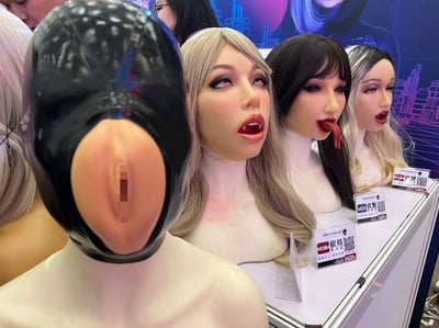 图片展示了一个展览中的几个人体模型头部。最左侧是一个黑色乳胶材质的面具，带有一个大的圆形开口。旁边是三个具有高度写实女性面容的模型头，分别拥有金色长发、黑色齐刘海长发和黑白渐变短发。这些模型妆容精致，表情各异，有的嘴巴微张，有的伸出舌头。每个模型前都有一个标有"DREAMASK"字样的价格标签。背景中可以看到模糊的人影和一个带有未来城市线条图案的展示板。