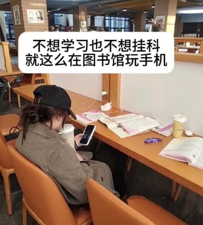 图片展示了一位学生在图书馆学习区的场景。学生戴着黑色帽子，背对着镜头，正在低头专注地玩手机。桌子上摆放着多本打开的课本、笔记本和两个饮料杯，但学生完全没有看学习资料，而是沉浸在手机屏幕中。图片上方有一行文字："不想学习也不想挂科 就这么在图书馆玩手机"，幽默地表达了许多学生既不想努力学习又害怕考试不及格，只能在图书馆假装学习实则玩手机的矛盾心理。