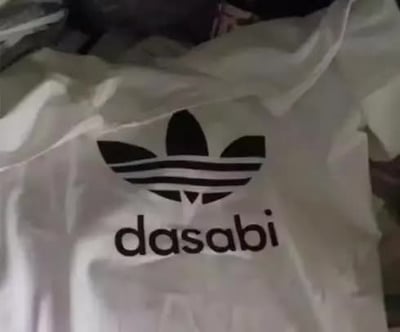 图片展示了一件白色T恤的特写，T恤正面印有黑色的三叶草图案（与运动品牌Adidas的经典标志高度相似），图案下方则是黑色的'dasabi'字样。整件T恤明显是模仿Adidas的山寨产品，通过篡改品牌名称制造出低俗或搞笑的效果。其中'dasabi'在部分方言中带有不礼貌含义，属于对知名品牌的恶搞。