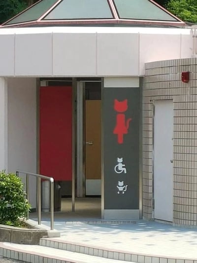 一张展示创意洗手间入口的照片，特色在于使用猫咪剪影代替了传统的人形图标。建筑具有现代风格，顶部为玻璃穹顶设计。主入口处有一个红色猫咪剪影标志，下方有两个白色猫咪图标 - 一个带有轮椅符号（无障碍设施），另一个为标准设计。入口处有金属扶手，显示这是一个考虑无障碍需求的公共设施。整体设计可爱有趣，将功能性指示牌与创意设计相结合。