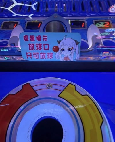 这是一张实拍的电玩城球类游戏机的局部照片，画面上方贴有浅蓝色背景的温馨提示标牌，标牌上用红色字体写着“温馨提示 放球口 只可放球”，文字旁搭配了一个有着浅白发、小角装饰的Q版动漫女孩，女孩做出手指触碰脸颊的动作，旁边还画了一颗红色小球，标牌正下方就是游戏机的圆形放球口，由于提示文案和配图的组合容易引发不雅歧义联想，被发布者吐槽配图糟糕。