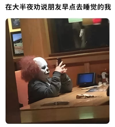 图片上方配有文字“在大半夜劝说朋友早点去睡觉的我”，下方是一名戴着白色小丑面具（面具上有黑色眼妆和红色鼻子）、顶着红色卷发的人，身穿深色外套，坐在木质桌子前，双手合十放在脸旁，侧身望向一边。背景可见木质墙壁、一个小屏幕和桌上的一些物品。通过小丑诡异的形象与“劝说睡觉”这一日常行为形成强烈反差，营造出幽默讽刺的效果。