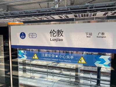 这是一张实拍的广州地铁伦教站1站台屏蔽门标识照片，标识清晰显示当前站点为伦教站（Lunjiao），位于F3层11号区域，下一站为广教站（Guangjiao）。标识下方配有中英文安全警示语“注意安全·小心夹手 / Caution! Watch Your Hands”，并搭配夹手警示的黄色图标，背景可看到地铁轨道，整体为公共交通站点的官方提示标识。