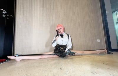 图中是一位粉色短发、佩戴眼镜的真人coser，身着白衬衫搭配黑色领结、黑色腰封短裙的制服风格cos服装，戴着黑色手套，正以一字马姿势坐在室内光滑地面上，左手扶着眼镜，右手持一把科幻风格的长剑，姿态帅气且展现出出色的柔韧性。背景是室内的浅灰色纹理墙面与深色门，场景简洁，重点突出coser的造型与姿态。