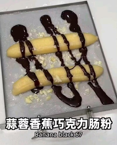 图片展示了一个银色托盘上放着两根类似肠粉的黄色长条状食物，表面淋有深色的巧克力酱，周围散落着切碎的蒜蓉。底部配有文字“蒜蓉香蕉巧克力肠粉 Banana black 67”，呈现出一种将香蕉、巧克力与蒜蓉这几种不常见搭配组合在一起的创意料理。