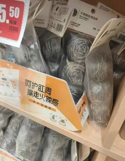 图片展示了超市货架上的一款商品，外包装为橙白配色的纸盒，盒上印有醒目的中文标语“呵护肛周 擦走火辣感”，暗示产品用于肛周护理。然而打开包装后可见内部为银色的不锈钢丝清洁球，这类清洁球通常用于厨房锅具清洁，与包装暗示的个人护理功能形成强烈反差，产生荒诞搞笑的效果。货架上还可见价格标签标注“5.50元/袋”，周围有多包同类商品堆叠。