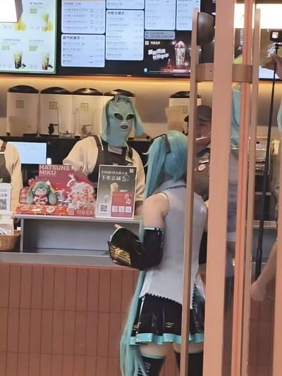 这是茶饮品牌与初音未来联名活动的门店现场，一位cos成初音未来的蓝发女生背对着镜头站在柜台前，柜台后的店员戴着与初音未来发色同色系的外星狗头面罩，趣味呼应coser的形象，柜台摆放着印有“HATSUNE MIKU”字样的联名宣传物料，还有“扫码领18元新人礼 下单立减5元”的优惠海报，旁边还有一位安保人员，整个场景充满玩梗趣味，是线下联动活动的整活实拍。