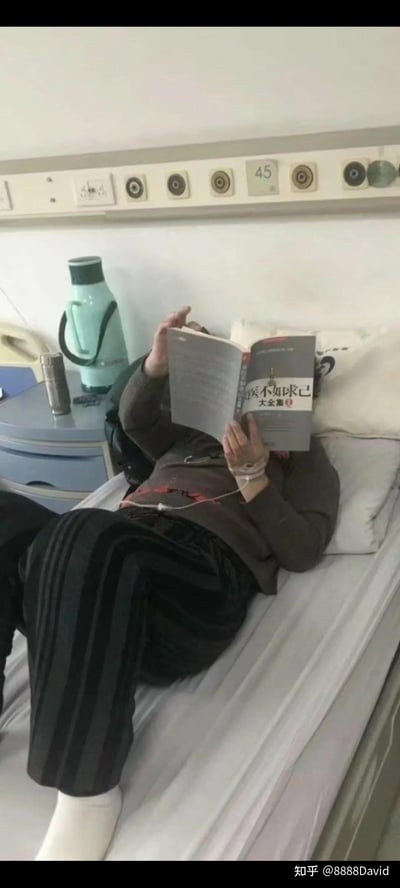 图中场景为医院病房，一名患者躺卧在病床上，上半身靠在枕头上，左手打着点滴，右手举着一本名为《买不如求己 大全集》的书遮挡面部阅读。患者穿着深色上衣和黑色条纹裤子，脚部盖着白色床单。病床旁的床头柜上放有绿色保温壶、银色保温杯及其他小物件，墙面上方有医疗设备接口和标有“45”的床位号标识。画面通过患者在生病输液时仍坚持阅读“求己”类书籍的场景，形成一种略带自嘲的幽默效果，暗示即便身处困境也试图通过自我帮助解决问题的心态。