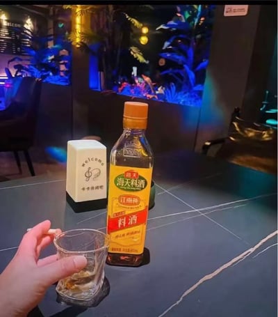 酒吧或休闲吧内的场景，深色桌面上摆放着一瓶海天料酒（黄色标签，烹饪用）、一个印有“Welcome 牛牛休闲吧”字样的白色方块摆件，以及一只手正拿着装有液体的玻璃杯。背景有蓝色灯光照射的植物，营造出温馨放松的酒吧氛围。图片通过将厨房常用的调料料酒置于酒吧场景中，形成一种反差萌，略带幽默效果。