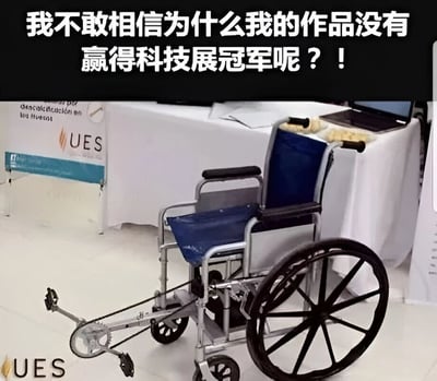图片展示了一把改装过的轮椅，轮椅主体为蓝色座椅和银色金属框架，前方装有特殊的机械装置。背景是一个铺着白色桌布的展台，上面放置着笔记本电脑等物品，旁边有带有"UES"标志的展板。图片上方配有文字："我不敢相信为什么我的作品没有赢得科技展冠军呢？！"，表达了对未能获奖的困惑和不解。