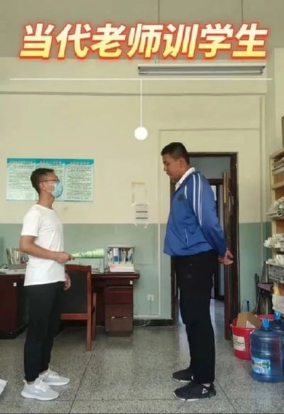 图片展示了一个办公室场景，房间内有两个人物。左侧是一位穿着白色T恤、戴着口罩的老师，手持一卷纸张，正面向右侧的学生。右侧学生穿着蓝色带白色条纹的运动外套和黑色裤子，双手背在身后，身体微微前倾，似乎在聆听老师讲话。背景中可见办公桌、椅子、墙上的宣传海报、悬挂的灯泡以及角落里的饮水机和水桶。图片顶部有橙色加粗文字“当代老师训学生”，整体呈现出老师正在教育学生的日常校园场景。