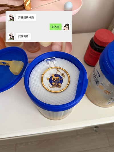 这张图的主体是摆放在白色台面上的开盖爱他美奶粉罐，奶粉罐的密封隔层上放置着金色手镯、带蝴蝶结吊坠的金项链等金饰，图片上方叠加了一段夫妻的聊天记录：男方先发消息说要开罐奶粉冲奶，女方回复询问对方在哪，男方称自己在厕所。结合画面内容可以推断，男方将金饰藏在奶粉罐内，开罐准备冲奶时临时去厕所，没来得及将藏的金饰收好，被女方发现，整件事充满生活化的幽默感。