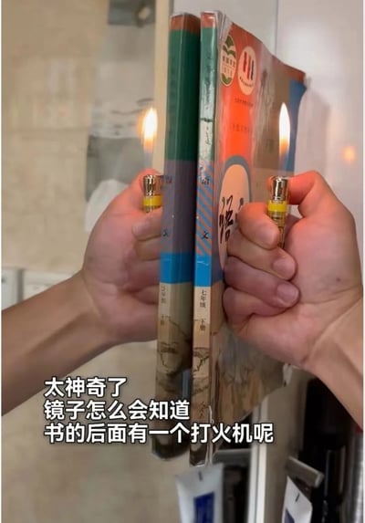 这是一张整活趣味图，拍摄场景为对着镜子拍摄：两只手分别举着一个点燃的打火机，放在两本七年级下册语文课本的左右两侧，镜子里反射出打火机的火焰，视觉上仿佛火焰分别位于书本两侧正对书本。图片下方配有故意装傻的文字，刻意忽略镜子会反射所有面前物体的基础常识，用一本正经疑惑的语气制造反差笑点。