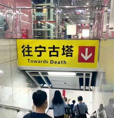 这是一张经过二次创作的地铁站指示牌照片，黄色指示牌上标注中文“往宁古塔”，下方的英文被恶搞成“Towards Death（走向死亡）”，右侧配有向下的红色箭头，画面下方能看到几位乘客正走下楼梯。
梗点说明：在清代历史中，宁古塔是流放重刑犯的偏远苦寒之地，在民间认知里是“有去无回”的象征，用“Towards Death”来翻译，精准贴合了这个历史意象，形成了荒诞搞笑的反差效果。