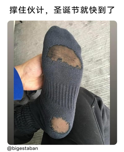 这是一张带有文字注解的真人实拍静态图，画面主体是一只手握着一只深蓝色的针织袜子，袜子的脚掌和脚跟位置都出现了明显的破洞，露出了皮肤；图片顶部配有文字“撑住伙计，圣诞节就快到了”，用诙谐的语气调侃自己穿着破袜子的窘迫处境，寄希望于圣诞节能获得新袜子或者改善这种小状况，传递出一种接地气的自嘲式生活幽默感，图片左下角带有@bigestaban的水印标识。