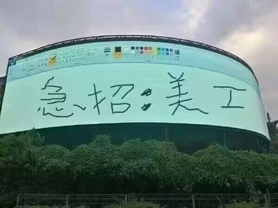 图片中展示了一个大型圆形广告牌，背景为浅色，顶部有彩色色卡及部分模糊文字。广告牌中央用黑色手写体醒目地写着“急招美工”四个大字，字体随意自然。广告牌前方是茂密的绿色灌木丛，天空呈现多云状态，整体画面形成鲜明反差：专业的大型广告载体与接地气的手写招聘信息结合，显得幽默又真实，带有一丝自嘲式的趣味。