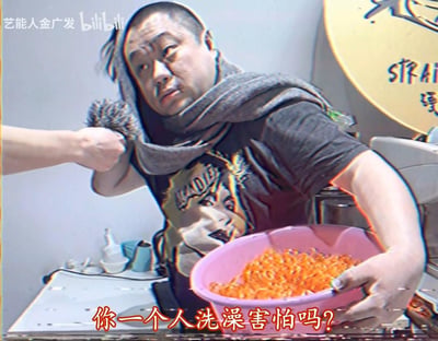 画面中一名男子身披灰色围巾，穿着印有图案的黑色T恤，左手端着一个装满橙色条状物体（疑似食物）的粉色水盆。另一只手从左侧伸出，拿着一个带毛球的工具靠近男子肩部。图片底部有醒目的红色文字'你一个人洗澡害怕吗？'，左上角有'艺能人金广发 bilibili'水印。
