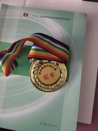 这张图片是一张实物拍摄的静态照片，主体是一枚带有彩色条纹绶带的金色冠军奖牌，奖牌中心印有红色的“冠军”字样，上方环绕着“萝莉魅魔榨精比赛”的文字，属于带有恶搞趣味的定制奖牌。奖牌放置在一本绿色封面的“十二五普通高等教育本科国家级规划教材”上，教材底部印有“高等教育出版社”的字样，背景还露出了一部分带横线的笔记本内页，正规教材与恶搞奖牌形成了鲜明的反差感，增添了趣味。