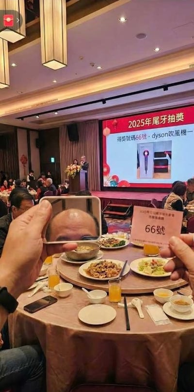 圖片展示了2025年尾牙抽獎現場，室內燈光溫暖，吊燈裝飾典雅，眾人圍坐餐桌享用宴席，桌上擺滿各式菜肴與飲品。舞台上有講台和花束，大屏幕顯示「2025年尾牙抽獎 得獎號碼66號 • dyson吹風機」字樣及戴森吹風機圖片。前景中一人雙手持物：左手拿手機拍攝，右手舉著寫有「66號」的粉色牌子，疑似中獎者記錄這一幸運時刻。