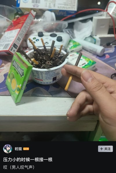 这是一张竖版社交媒体截图，主体是一张桌面实拍图。画面中央的白色小碗里装满压碎的黑色饼干屑，许多巧克力涂层饼干棒插在其中，浅色饼干末端露出，远看很像插在烟灰缸里的烟头。右侧有一只手用食指和拇指捏住一根饼干棒，姿势高度模仿夹香烟。碗的左侧斜放着一盒绿色包装的绿箭口香糖，背景还能看到白色 Xbox 风格游戏手柄、纸巾、充电头、充电线和其他零食包装，整体是普通宿舍或卧室桌面环境。截图底部显示发帖用户为“輕朧”，等级为 Lv.15，配文写着“压力小的时候一根接一根 哎（男人叹气声）”。图片利用饼干屑和饼干棒模仿烟灰缸与烟头，制造“抽烟解压”的第一眼错觉，实际表现的是不停吃零食，带有自嘲、吐槽和搞笑效果。