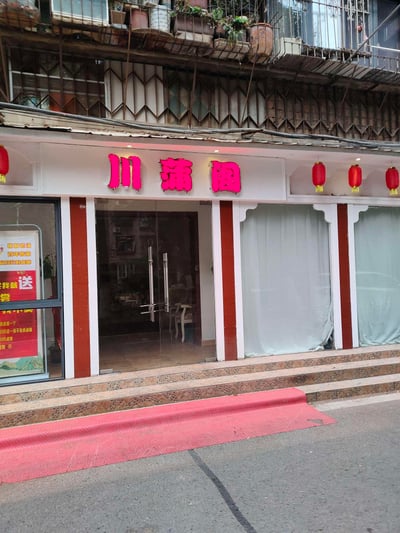 照片展示了一家名为“川蒲阁”的餐馆门面。门面上方有红色霓虹灯招牌“川蒲阁”，两侧悬挂着四个红色灯笼，营造中式传统氛围。正门为玻璃双开门，门内可见部分店内陈设；门两侧是带有白色窗帘的窗户，窗框为红白配色。入口处有三级台阶，台阶前铺着红色防滑垫。左侧玻璃窗上贴有红色促销海报，部分文字显示“我就送”“学”等字样。建筑二楼阳台摆放着多个花盆，栏杆处可见老旧的金属结构，整体呈现出市井生活中传统餐馆的典型外观。