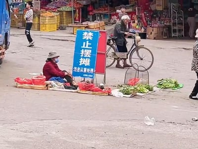 街头场景中，一位戴着帽子和口罩的老人坐在地上，面前摆着蔬菜摊位，摊位旁立着一个蓝色的'禁止摆摊 违者从重处理'的警示牌。背景中有其他行人和停放的自行车，远处是水果摊位和黄色的货箱，展现了日常街头的矛盾画面。