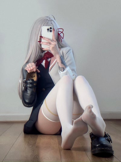 一位银发少女穿着校园风格的制服自拍，她身着白色西装外套、黑色背心和红色领结，搭配白色过膝长袜，袜子顶部有金色环状装饰。少女左手拿着手机挡住面部，右手握着一只黑色的玛丽珍鞋，手臂上有明显的纹身图案。她坐在木地板上，双腿交叉，背景是纯白色的墙壁，整体呈现出一种日系校园的穿搭风格。