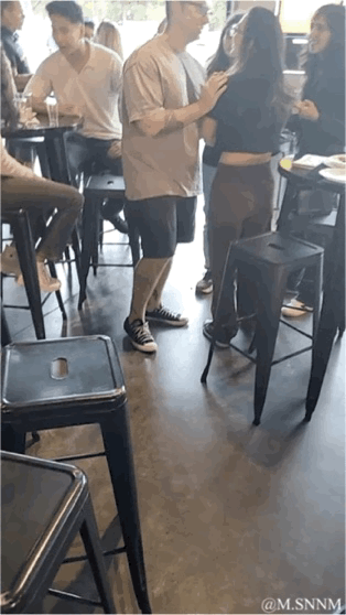 在一个热闹的室内餐吧/酒吧场所，戴眼镜的男子正和身边的女生开心互动，此时画面下方突然伸出一只手做出握手邀请的手势。男子见状立刻转身，伸手准备回应握手，结果却发现并没有人在对面，他一脸疑惑地转回身，手臂上的纹身也露了出来。整个场景是一个利用视觉差设计的整蛊恶作剧，通过虚假的握手邀请制造出搞笑的反转效果，现场氛围轻松欢乐。