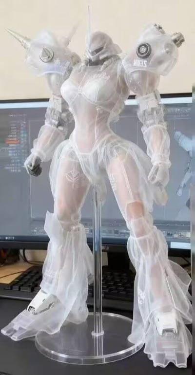 图片展示了一个白色半透明机甲风格人形手办，整体采用透明塑料材质，呈现出独特的科技感。手办具有明显的女性人体轮廓，同时融合了丰富的机械细节：肩部装有带尖刺的装甲组件，手臂配备圆形关节和印有“NEXUS”字样的护肩，腿部覆盖透明裙甲状结构，脚部设计有机械靴。手办站立在透明支架上，背景为两台电脑显示器，屏幕上显示着3D建模软件界面，推测该手办可能是基于屏幕中的设计文件制作的原型或定制模型。