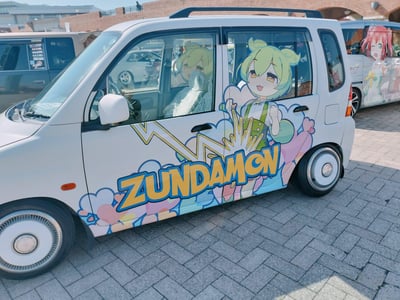 这是一辆以VOICEROID虚拟主播纯位数（Zundamon）为主题的日式K-car痛车，车身侧面绘制了绿发的Zundamon握拳欢呼的可爱形象，搭配彩色爱心与闪电装饰图案，车门下方印有醒目的黄色描蓝边的“ZUNDAMON”字样。车内车窗也张贴了同款角色的贴纸，背景中可见其他二次元主题痛车，显示这是一场痛车聚会或ACG相关活动的现场，整体风格充满活泼可爱的二次元气息。