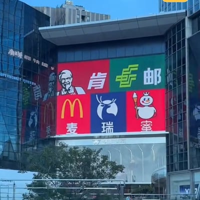 图片展示了一个大型户外广告牌，上面创意性地组合了五个知名品牌的标志和名称。广告牌采用2x3的网格布局：左上角是肯德基创始人桑德斯上校的肖像，右上角是中国邮政的绿色标志和文字，左下角是麦当劳的黄色M标志和'麦'字，中间是瑞幸咖啡的蓝色鹿头标志和'瑞'字，右下角是蜜雪冰城的雪人吉祥物和'蜜'字。背景是现代化的玻璃幕墙建筑，底部可见'世茂广场'字样。这种将快餐、咖啡品牌与邮政服务品牌并置的组合方式产生了幽默有趣的视觉效果。