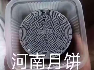 一张创意幽默梗图，画面主体是一个圆形井盖被放置在透明塑料食品餐盒中。井盖表面有交错的菱形网格纹路，中心刻有“市政”等字样及相关标识，整体质感粗糙厚重。图片底部叠加了醒目的白色大字“河南月饼”，通过将井盖的外形、纹路与传统月饼进行夸张联想，制造出“硬核月饼”的诙谐视觉效果，调侃其形态酷似月饼的趣味对比。