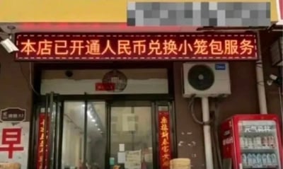 这是一张线下小笼包餐饮店的实拍照片，店铺门头上方的红色发光电子招牌上写有“本店已开通人民币兑换小笼包服务”的字样，实质上就是普通的付费购买小笼包的消费行为，商家刻意用“兑换服务”的官方化表述玩梗，制造出荒诞的幽默感。画面中还能看到店铺的玻璃推拉门、门两侧的红色春联、墙面的空调外机、右侧的饮料冷藏柜，以及左侧带有“早”字的早餐标识，整体是常见的市井早餐店的样貌。