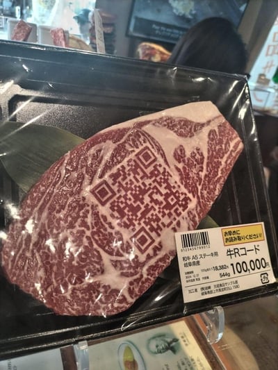 这是一块包装好的日本岐阜县产A5级和牛牛排，最具创意的是牛排的大理石花纹（脂肪与瘦肉的分布）恰好形成了类似QR二维码的图案，完美结合了食品溯源的概念。标签显示该牛排售价高达100,000日元，每100克单价18,382日元，重量544克，赏味期限标注为3004年12月31日（属于夸张设定，突出概念性），是用于制作牛排的顶级和牛。这是创意营销内容，通过新奇的视觉设计宣传和牛的高端定位与可溯源特性，视觉冲击力极强，展现了高端食材的独特创意呈现。