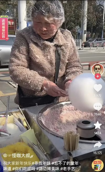 这张图展示了在中国街头制作棉花糖的阿姨，阳光下阿姨正熟练地用机器绕制棉花糖，旁边摆有各色糖丝和竹签。界面为抖音短视频，点赞评论数量众多，说明此内容受网友喜爱。画面透出浓浓的民间市井气息，主要围绕童年路边摊美食和传统手艺怀旧氛围。没有明显谐音梗或双关，但极具烟火气和人情味，适合激发关于年味与童年的共鸣。