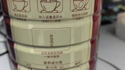 图片展示了某产品（疑似食品或冲调类饮品）的标签特写，标签上印有使用说明和防伪提示。具体内容包括：取本品、加入适量温开水（约85℃）冲泡、搅拌均匀后即可食用的使用步骤，以及“防伪码处：此处未贴防伪码 一律为假货”的防伪警告。背景为模糊的桌面环境，整体拍摄角度为竖版近距离拍摄，标签文字清晰可见。