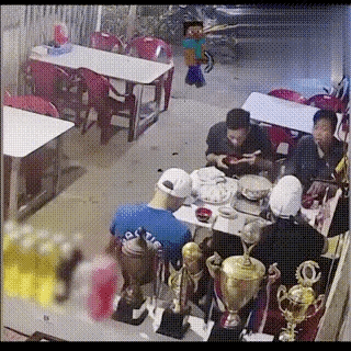 这是一段搞笑的动态GIF，内容展示了现实里的一场聚餐：几名年轻人围坐在摆着食物和奖杯的餐桌旁吃饭，突然游戏《我的世界》中的角色史蒂夫出现在画面后方，紧接着餐桌区域出现类似爆炸的特效，聚餐的人们瞬间惊慌失措，纷纷逃离现场，最后只剩下一人留在原地。视频通过将游戏角色与真实监控场景合成，制造出荒诞无厘头的反差搞笑效果。