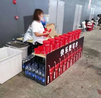 这是一张实拍的线下活动饮品摊位照片，摊位正面立着深棕色招牌，招牌上写着白色大字“快乐肥宅水”，招牌上方的台面上整齐摆放了一排带盖的红色可口可乐杯子。一名穿着白色短袖、戴着口罩的短发女士站在摊位后方，正在整理摆放红色杯子。摊位招牌后方的支撑位置，摆放了多瓶大瓶装百事可乐作为支撑，形成了可口可乐摊位用竞品百事可乐当底座的趣味反差场景。摊位左侧放置了白色保温箱和容器，背景是灰色墙面的活动场馆通道，远处还能看到其他桌椅和活动人员。