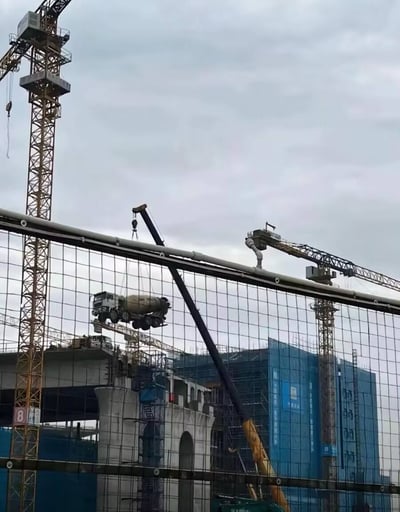 这张图片展示了一个繁忙的建筑工地场景。画面中央有一台起重机正吊起一辆完整的混凝土搅拌车，悬停在半空之中。前景是建筑工地常见的安全防护网，背景中可以看到多台塔式起重机和正在建设中的建筑物。天空呈现出阴天的灰白色调，整个场景展现了建筑工程中不常见的大型设备吊装作业，给人一种视觉上的震撼感。