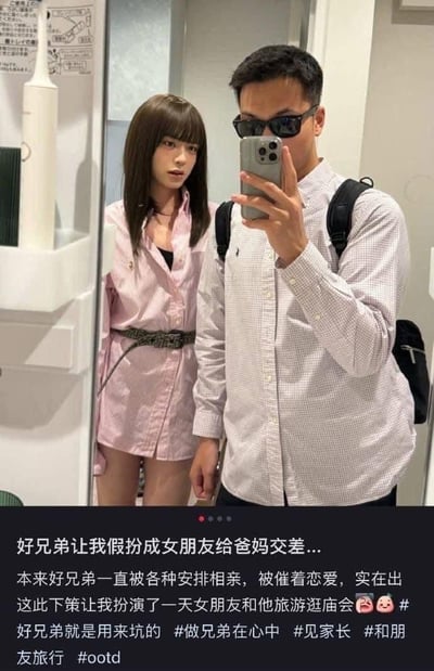 图片是一张社交平台的图文截图，画面主体是镜子前的两个人：右侧是一位戴着黑框墨镜、穿格子衬衫背双肩包的男生，正拿着iPhone手机对着镜子自拍；左侧的“女生”实则是男生假扮的，留着齐刘海棕色长发，穿着粉色长款衬衫当连衣裙，搭配腰带，露出腿部，表情带着无奈和尴尬。下方配文讲述了故事背景：这位假扮女友的男生，因为好兄弟一直被家人安排相亲、催着恋爱，无奈之下出此下策，让自己假扮成他的女朋友，一起旅游逛庙会，帮兄弟给爸妈交差，还附带了多个相关话题标签，整体呈现出兄弟间搞笑又无奈的趣事。