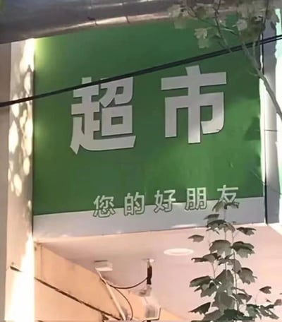 一张超市招牌的照片，绿色背景上有白色大字'超市'，下方有较小文字'您的好朋友'。其中'朋'字使用了不规范的写法，看起来像是一个错别字。照片中可以看到招牌周围的建筑结构和一些植物枝叶。