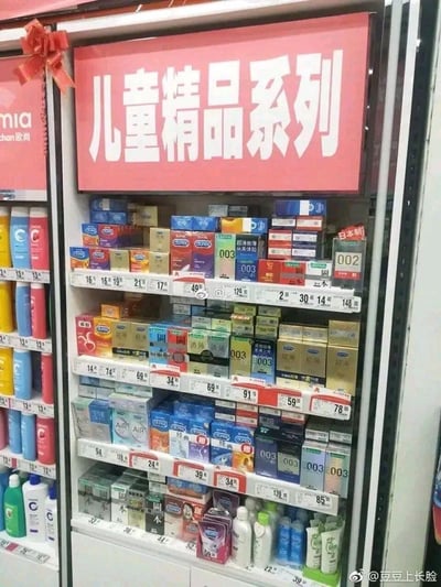 这是一张超市货架的实拍照片，货架上方悬挂着醒目的红色标牌，上面写着“儿童精品系列”，但货架的多层空间里却密密麻麻摆满了各类品牌的避孕套产品，包括杜蕾斯、杰士邦等知名计生用品品牌，不同包装、价位的避孕套整齐陈列，与“儿童精品”的标牌形成极其强烈的反差感，制造出荒诞又搞笑的错位效果，显然是超市商品摆放失误或是故意的整活行为，这种场景错位形成了极具笑点的梗图内容。