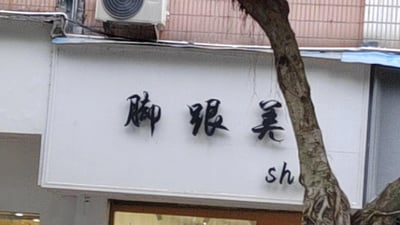图片展示了一个街头店铺的白色招牌，上面用黑色书法字体写着'脚跟美'，右下角可见部分英文字母'sh'。招牌背景为居民楼外墙，上方装有空调外机，右侧有一棵树干斜穿画面，整体呈现出真实的城市街景。招牌名称'脚跟美'字面意思为'脚跟美丽'，推测可能是提供足部护理、美甲等服务的店铺。